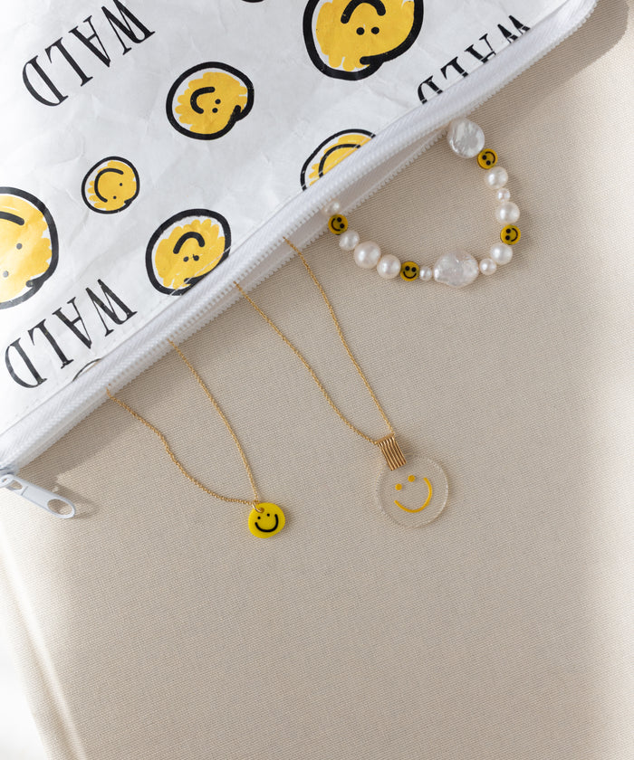 The WALD Berlin Smilie Yellow Necklace is arranged alongside a beaded bracelet and a clear hand-painted pendant with smiley faces, next to a white zipper pouch featuring a matching smiley design, all displayed on a beige surface.