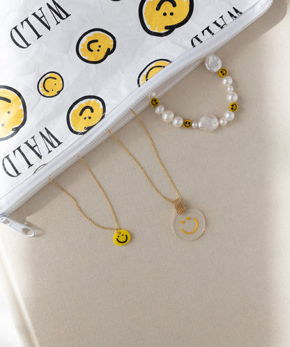 The WALD Berlin Smilie Yellow Necklace is arranged alongside a beaded bracelet and a clear hand-painted pendant with smiley faces, next to a white zipper pouch featuring a matching smiley design, all displayed on a beige surface.