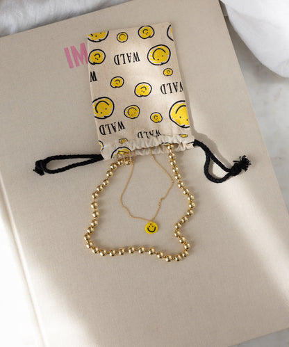 A beige surface displays a drawstring pouch adorned with smiley faces, a gold beaded necklace, and the WALD Berlin Smilie Yellow Necklace—a gold-plated accessory featuring a hand-painted pendant.