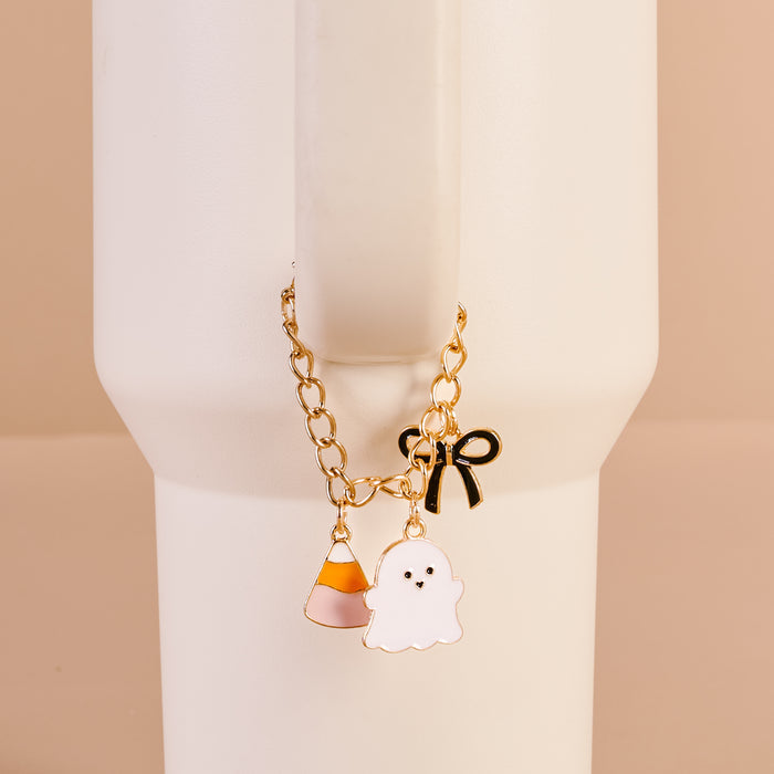 The Darling Effect's Spooky Sweet Tumbler Charm—a gold chain with candy corn, ghost, and black bow—hangs from a tall white cup's handle against a beige background, perfect as a Halloween accessory or bracelet.