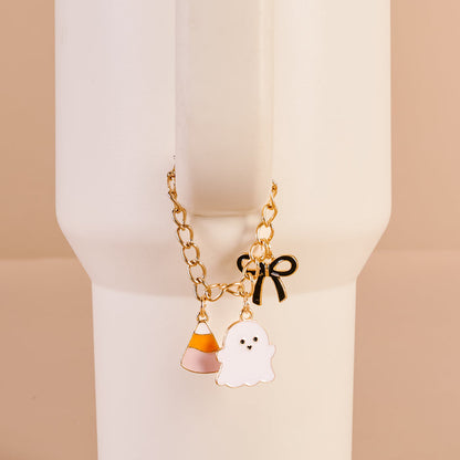 The Darling Effect's Spooky Sweet Tumbler Charm—a gold chain with candy corn, ghost, and black bow—hangs from a tall white cup's handle against a beige background, perfect as a Halloween accessory or bracelet.