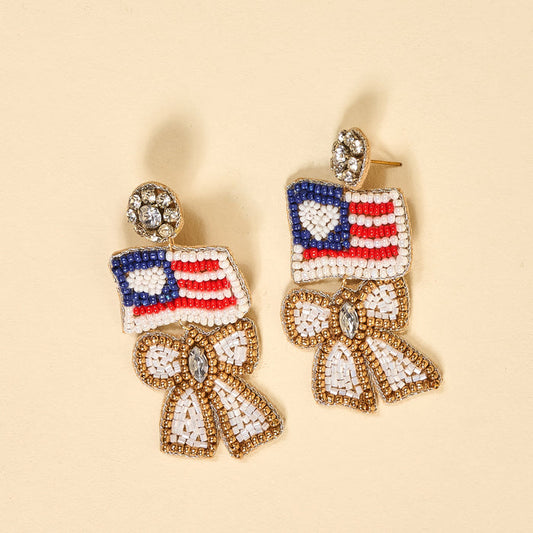 The Darling Effect’s Star Spangled Bow Beaded Earrings feature American flag-inspired red, white, and blue beads above rhinestone-studded bows on beige—a bold patriotic jewelry statement.