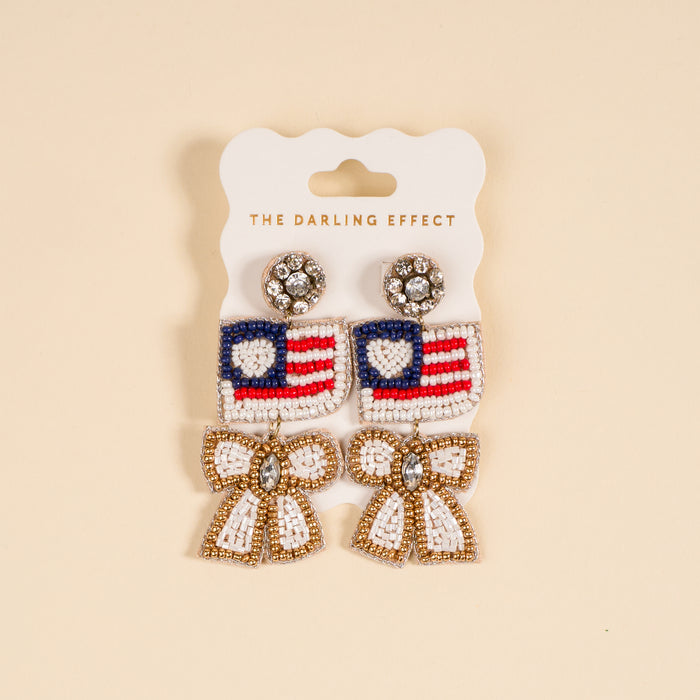 The Darling Effect’s Star Spangled Bow Beaded Earrings feature hand-beaded American flag hearts, accented with dangling white and gold bows and sparkling rhinestones, presented on a branded card.