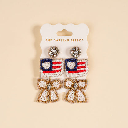 The Darling Effect’s Star Spangled Bow Beaded Earrings feature hand-beaded American flag hearts, accented with dangling white and gold bows and sparkling rhinestones, presented on a branded card.