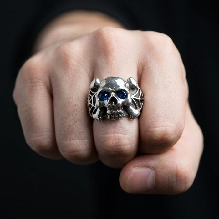 Silver skull and crossbones ring with blue eyes and spider web details worn on a man's fist.