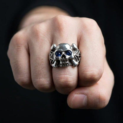 Silver skull and crossbones ring with blue eyes and spider web details worn on a man's fist.