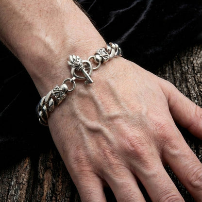 A man's wrist wearing a chunky sterling silver curb chain bracelet featuring an ornate gothic heart and key toggle clasp, set against a rustic wood and dark velvet background.