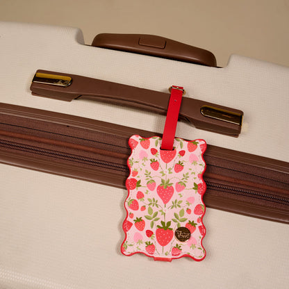 The Darling Effect’s Strawberry Fields Jetsetter Luggage Tag—a pink tag with a privacy flap, red strawberries, and a wavy border—adds charm to any white suitcase with brown zippers and handle.
