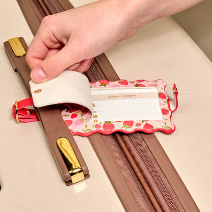 A hand lifts the privacy flap of The Darling Effect’s Strawberry Fields Jetsetter Luggage Tag on a beige suitcase, revealing an information card inside this charming travel accessory.