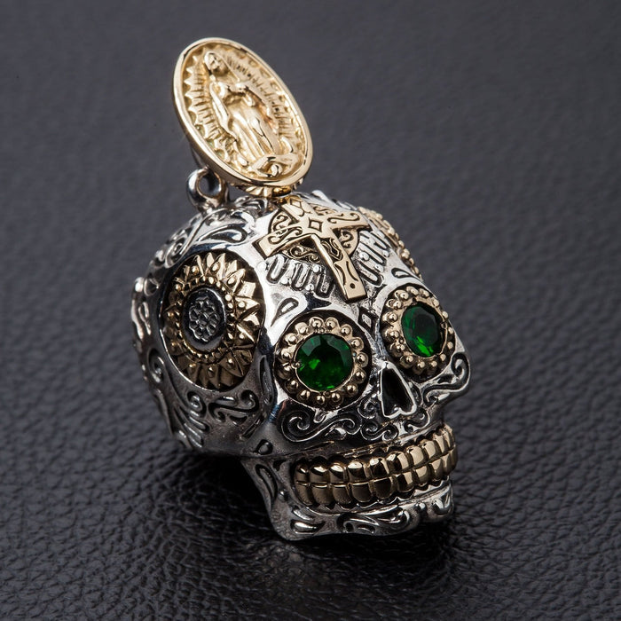 A handcrafted sterling silver sugar skull pendant with green gemstone eyes and a gold Virgin Mary bail, shown on a black background.