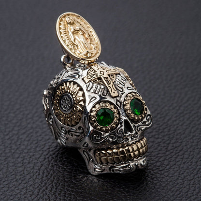 A handcrafted sterling silver sugar skull pendant with green gemstone eyes and a gold Virgin Mary bail, shown on a black background.