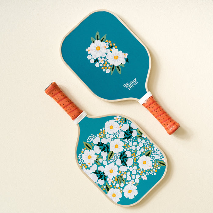 Two Sweet Meadow Green Pickleball Paddles by The Darling Effect, featuring blue floral designs and orange handles, rest on a light background—each showcasing white and yellow flowers in different-sized clusters. Perfect for any pickleball enthusiast.