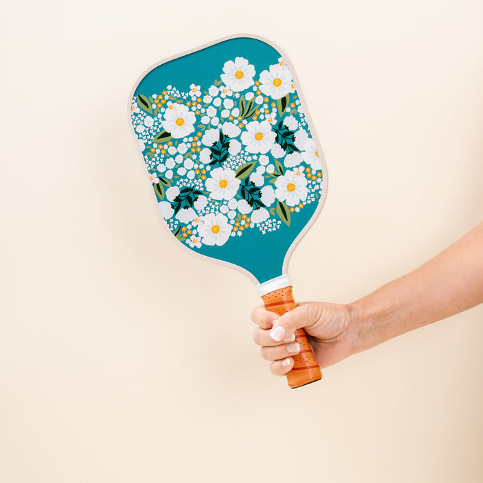 A hand holds The Darling Effect's Sweet Meadow Green Pickleball Paddle, featuring a teal floral design on a light beige background—ideal for pickleball enthusiasts.