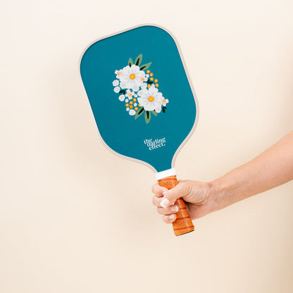 A hand holds The Darling Effect's Sweet Meadow Green Pickleball Paddle, featuring a teal floral design and logo—perfect for any pickleball enthusiast.
