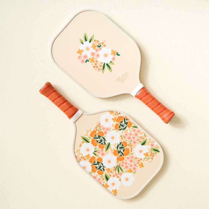 Two Sweet Meadow Tan Pickleball Paddles by The Darling Effect, featuring floral white, orange, and green designs with orange handles, are displayed side by side on a light-colored surface—perfect for any pickleball enthusiast.