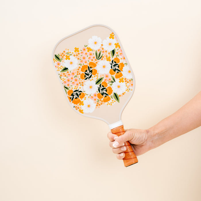 A hand holds The Darling Effect's Sweet Meadow Tan Pickleball Paddle, featuring an orange, yellow, and white floral pattern that pops against a plain cream background—ideal for any pickleball enthusiast.