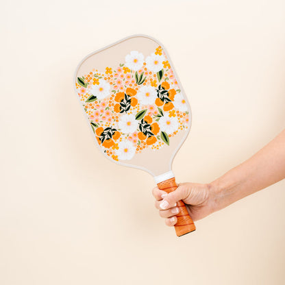 A hand holds The Darling Effect's Sweet Meadow Tan Pickleball Paddle, featuring an orange, yellow, and white floral pattern that pops against a plain cream background—ideal for any pickleball enthusiast.