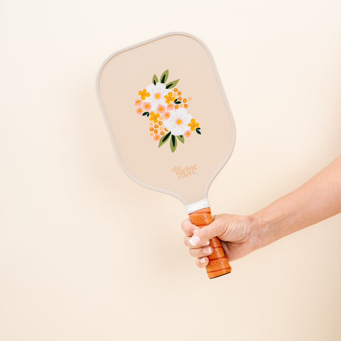 A hand holds the Sweet Meadow Tan Pickleball Paddle by The Darling Effect, featuring orange and white flowers, green leaves, and “the darling effect” text—a stylish choice for any pickleball enthusiast.
