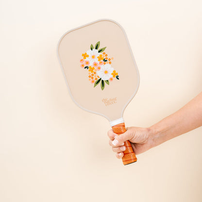 A hand holds the Sweet Meadow Tan Pickleball Paddle by The Darling Effect, featuring orange and white flowers, green leaves, and “the darling effect” text—a stylish choice for any pickleball enthusiast.