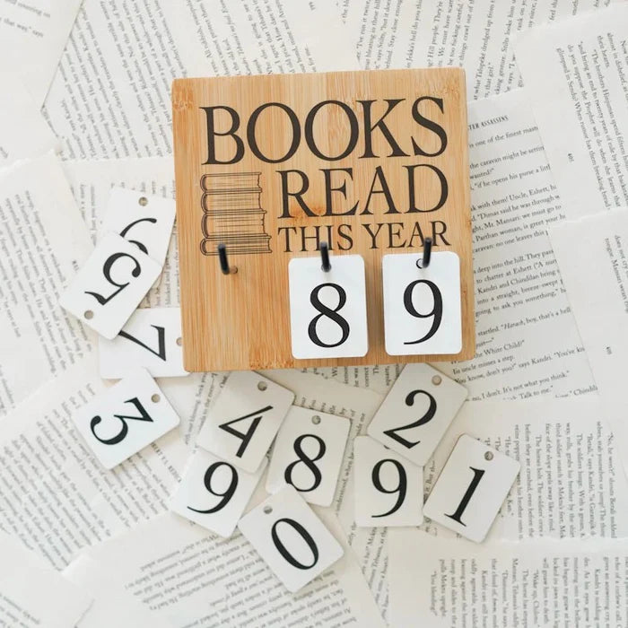 This image shows a wooden sign with the text "BOOKS READ THIS YEAR" and two hanging white tags displaying the numbers "8" and "9". The sign is placed on top of a collection of white tags with various numbers and letters, which appear to be part of a larger set. The background consists of scattered pieces of paper with printed text, creating a textured and somewhat chaotic appearance. The overall theme suggests a focus on reading and tracking the number of books read within a year.