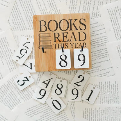 This image shows a wooden sign with the text "BOOKS READ THIS YEAR" and two hanging white tags displaying the numbers "8" and "9". The sign is placed on top of a collection of white tags with various numbers and letters, which appear to be part of a larger set. The background consists of scattered pieces of paper with printed text, creating a textured and somewhat chaotic appearance. The overall theme suggests a focus on reading and tracking the number of books read within a year.