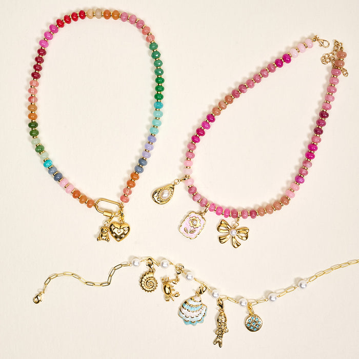 Three colorful beaded necklaces from The Darling Effect are displayed on a beige background, featuring gold charms like hearts and bows. One necklace showcases pearls and the Teddy Bear Jewelry Charm Gold from the charm bar collection.