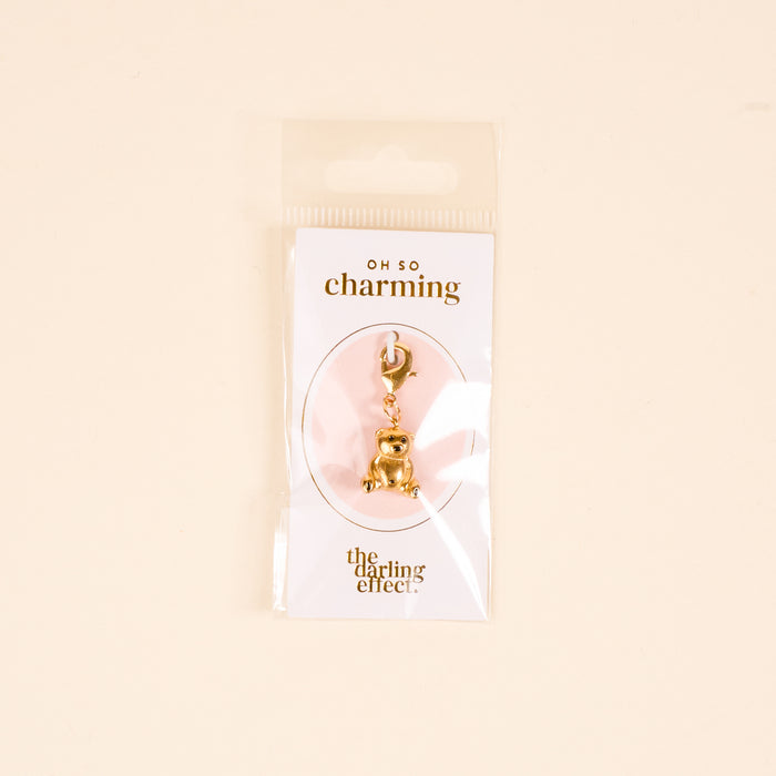 A gold teddy bear jewelry charm by The Darling Effect is attached to a card labeled OH SO charming and packaged with the card and charm bar in a clear plastic bag against a beige background.