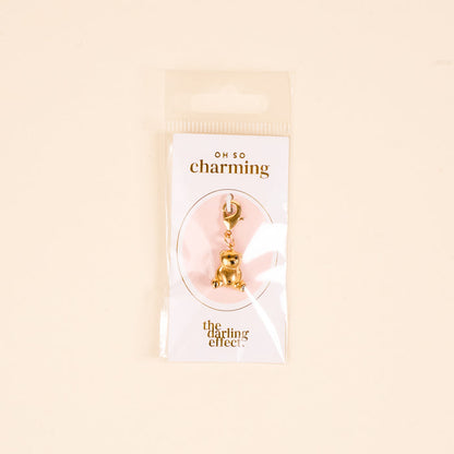 A gold teddy bear jewelry charm by The Darling Effect is attached to a card labeled OH SO charming and packaged with the card and charm bar in a clear plastic bag against a beige background.