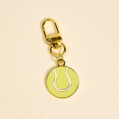 The Darling Effect's Tennis Ball Pawfect Collar Charm features a gold keychain with a yellow and white tennis ball—ideal for adding playful style to your keys or as a trendy accessory for your pet’s collar.