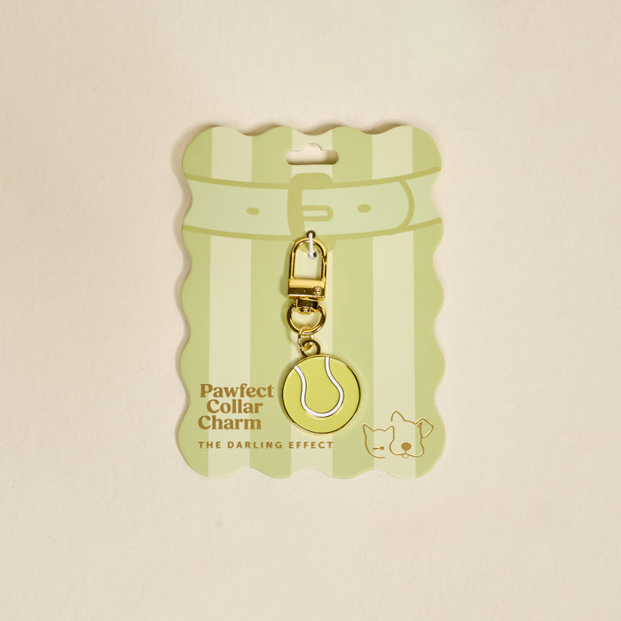 The Darling Effect's Tennis Ball Pawfect Collar Charm features a gold and yellow tennis ball design with a clasp, cute dog and cat illustrations, and comes on striped Pawfect Collar Charm packaging for playful pet style.