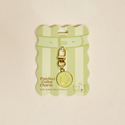 The Darling Effect's Tennis Ball Pawfect Collar Charm features a gold and yellow tennis ball design with a clasp, cute dog and cat illustrations, and comes on striped Pawfect Collar Charm packaging for playful pet style.