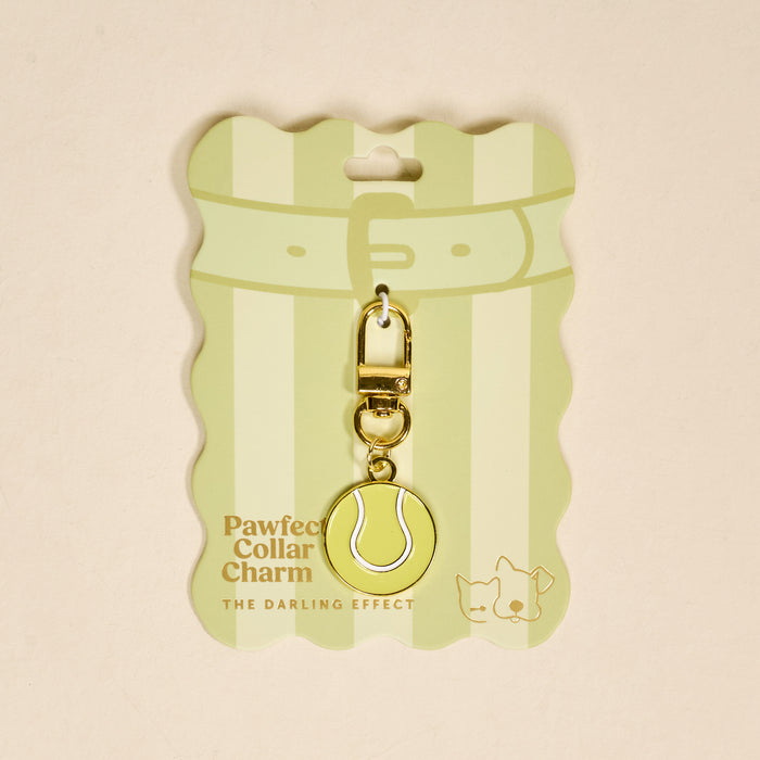 The Darling Effect's Tennis Ball Pawfect Collar Charm is a gold, tennis ball-shaped charm with clasp, displayed on striped green packaging featuring cute cat and dog illustrations—perfect for playful pet style lovers.