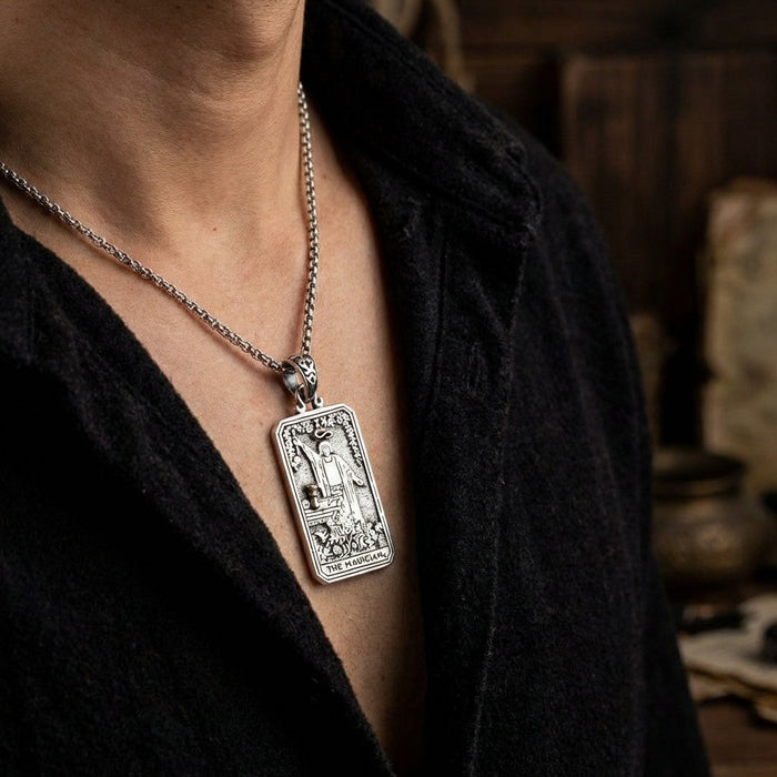 Close-up of a man wearing a detailed silver Magician tarot card pendant on a chain necklace, styled with an open black shirt against a moody, rustic background.