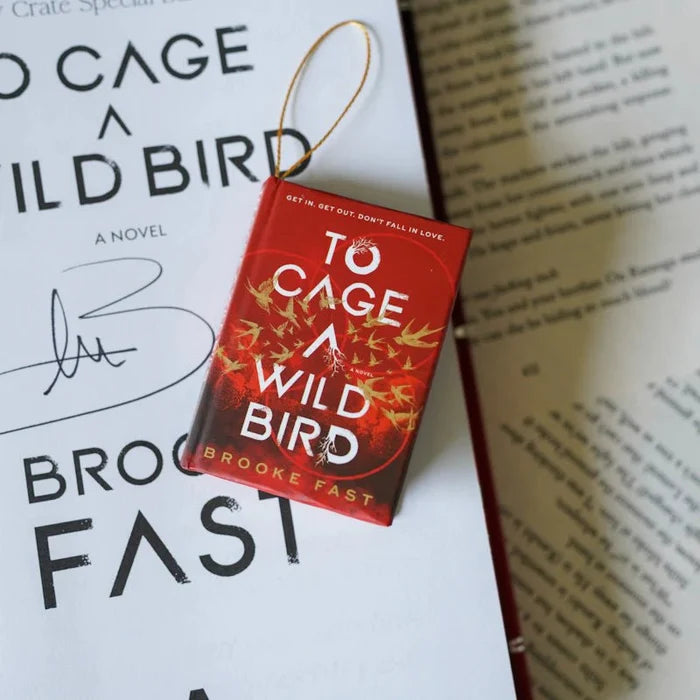 To Cage a Wild Bird by Brooke Fast LitJoy Crate exclusive mini book ornament with gold ribbon loop, featuring a red cover with golden bird illustrations and the tagline "Get in. Get out. Don't fall in love.", resting on the full-size signed hardcover edition next to an open book