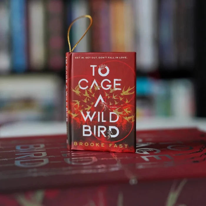 To Cage a Wild Bird by Brooke Fast LitJoy Crate exclusive mini book ornament standing upright on a stack of red hardcover books, with a gold twisted cord loop at the top, red cover featuring golden bird illustrations and tagline "Get in. Get out. Don't fall in love.", with a blurred colorful bookshelf in the background