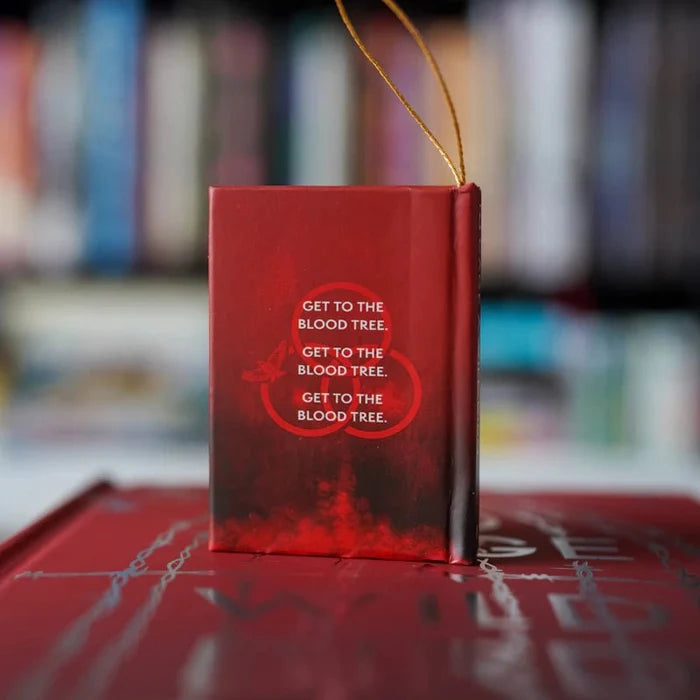 Back cover of the To Cage a Wild Bird LitJoy Crate exclusive mini book ornament, showing a deep red design with the repeated quote "Get to the Blood Tree." and a circular illustrated motif, gold twisted cord loop at the top, resting on a stack of red hardcover editions with blurred bookshelf background