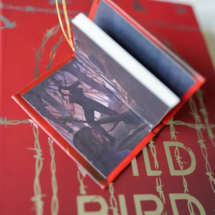 To Cage a Wild Bird LitJoy Crate exclusive mini book ornament opened like a locket, revealing a character art print inside showing a dark-clothed figure in a moody forest scene, with the red barbed wire edition cover visible and gold ribbon loop at the top, resting on the hardcover barbed wire edition