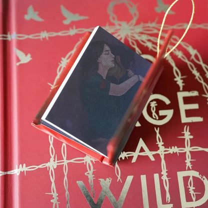 To Cage a Wild Bird LitJoy Crate exclusive mini book ornament open at an angle, revealing interior character art of a tender romantic couple embracing — a dark-haired woman with eyes closed leaning into a man's arms — with the red barbed wire LitJoy edition hardcover visible in the background and gold cord loop at top