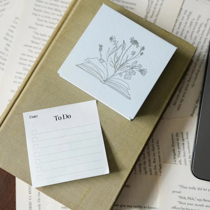 To Do Sticky Notes pad close-up from LitJoy Winter Romantasy TBR Box — white notepad with date and checkbox fields beside botanical flower illustration card, on open book