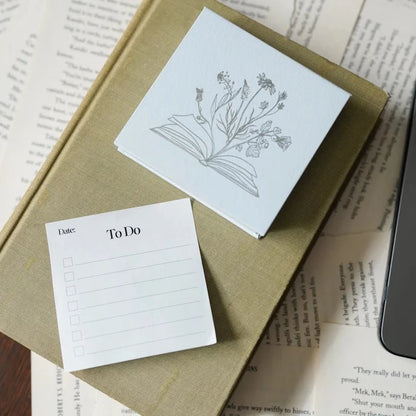 To Do Sticky Notes pad close-up from LitJoy Winter Romantasy TBR Box — white notepad with date and checkbox fields beside botanical flower illustration card, on open book