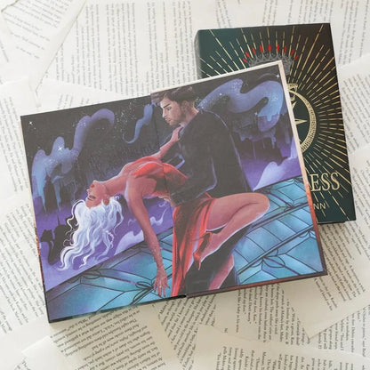This image shows an open book with a fantastical illustration on the page. The illustration depicts a man and a woman dancing in an embrace, set against a backdrop of a night sky with stars and a cityscape. The book is placed on top of several sheets of paper with printed text. The book itself has a decorative cover with intricate designs and the word "ESS" prominently displayed on it