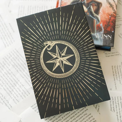 This image shows a black rectangular object with a golden design featuring a star and a snake forming a circle, placed on top of several sheets of paper. The object appears to be a book or a box with a striking and intricate design. Behind it, partially visible, is another book with the name "WINN" on the cover, suggesting it might be authored by someone named Winn. The papers beneath contain various text snippets, some of which are partially obscured by the object on top.