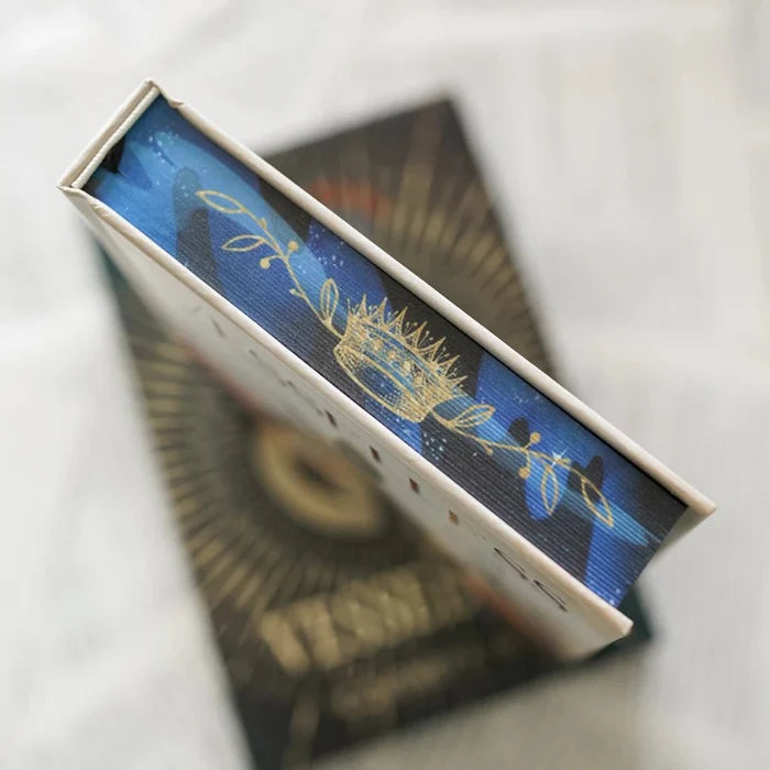 This image shows a close-up view of a book with an intricate design on its top page edge. The page edge features a blue background with a golden crown and floral patterns. The book appears to be placed on top of another object, which has some text and a golden design. The text on the lower object is partially visible and includes repeated characters and words. The overall scene suggests a focus on the detailed artwork and design elements of the book cover.