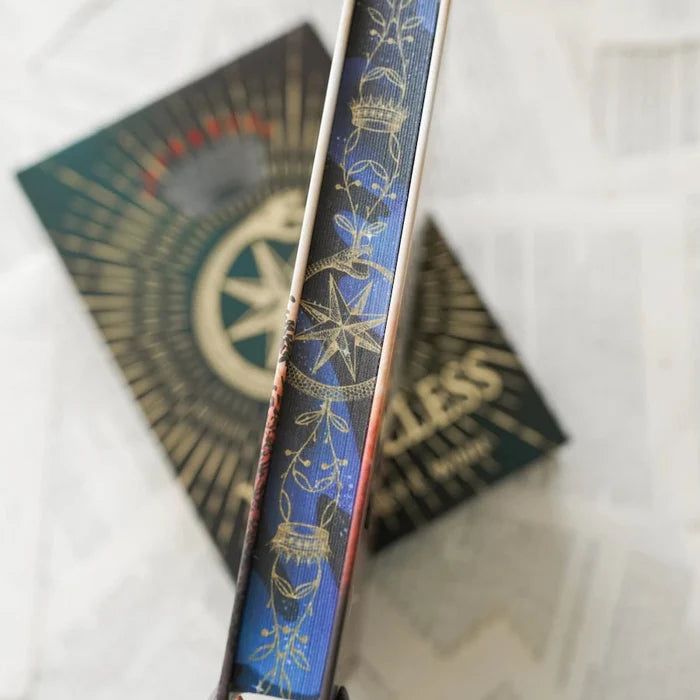 This image shows a close-up view of a book page edges with intricate designs. The page edges features a blue background with gold and white decorative elements, including a crown, a compass rose, and floral patterns. The text "LESS" is visible on the spine, repeated multiple times. The book appears to be placed on a textured surface, possibly fabric or paper, which provides a contrasting background to the detailed artwork on the spine.