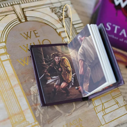 We Who Will Die LitJoy mini book ornament opened to reveal a full-color interior illustration of armored warriors in battle, resting on the beige gold LitJoy exclusive cover with the purple edition visible behind it