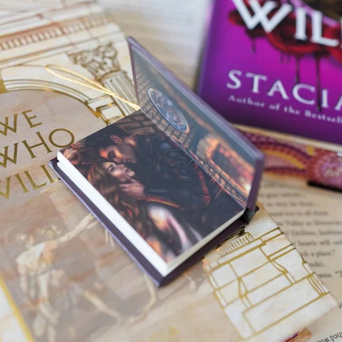 We Who Will Die LitJoy mini book ornament opened to a full-color interior illustration of a red-haired woman in a dramatic architectural scene, resting on the beige gold LitJoy exclusive cover with the purple edition in the background