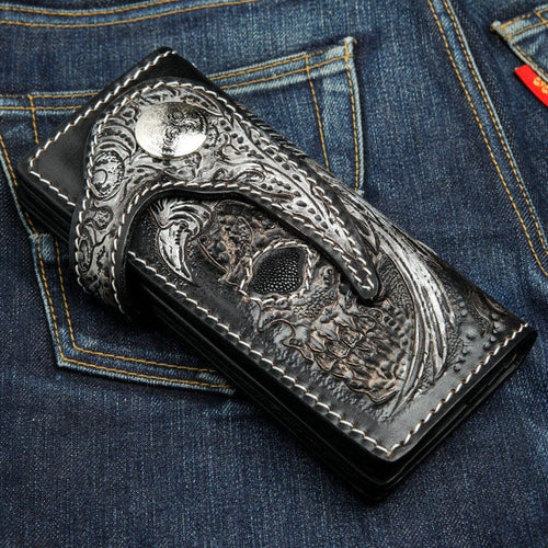 Hand-carved winged skull biker wallet front view showing detailed leather carving and black stingray inlay on denim background