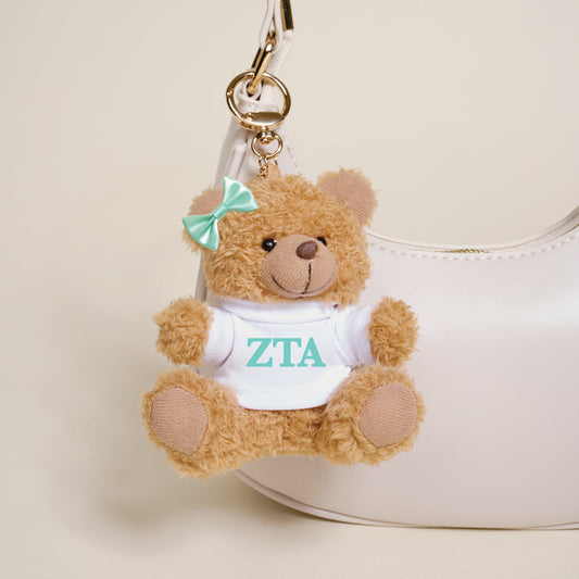 The Darling Effect's Zeta Tau Alpha Teddy Bear Bag Charm features a small brown bear with a green bow and white “ZTA” shirt—adorable on any white handbag and perfect for Bid Day baskets or as an everyday sorority accessory.