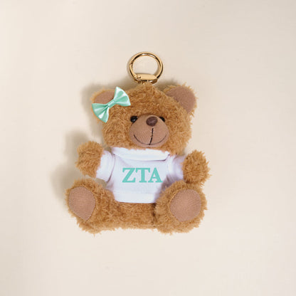 The Darling Effect Zeta Tau Alpha Teddy Bear Bag Charm is a small brown plush keychain with a teal bow and white shirt with teal ZTA letters—perfect for Bid Day baskets or as a cute bag accessory. Set against a light beige background.